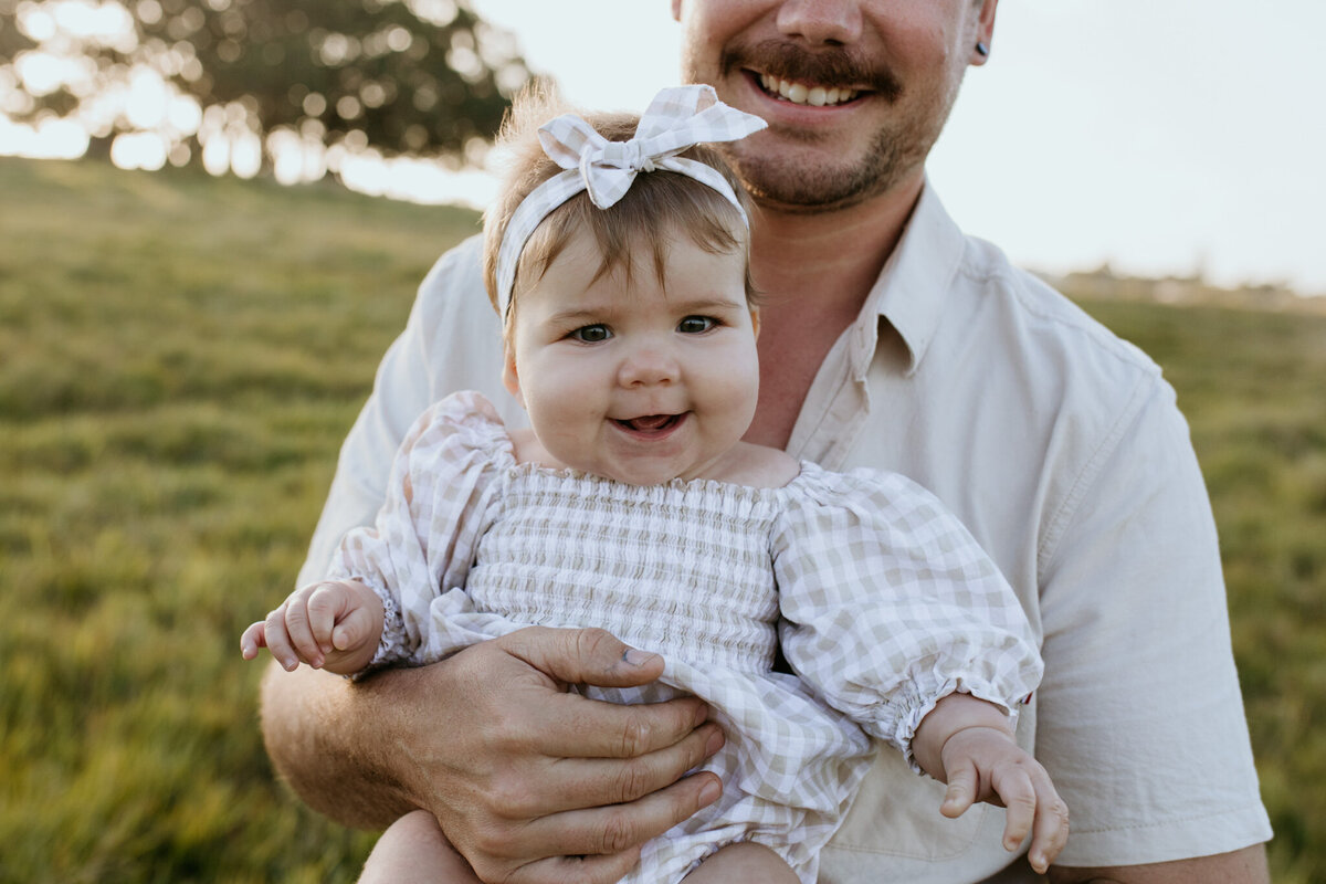 Six-month-old baby giggles broadly while her dad holds her, his hand resting on her belly.