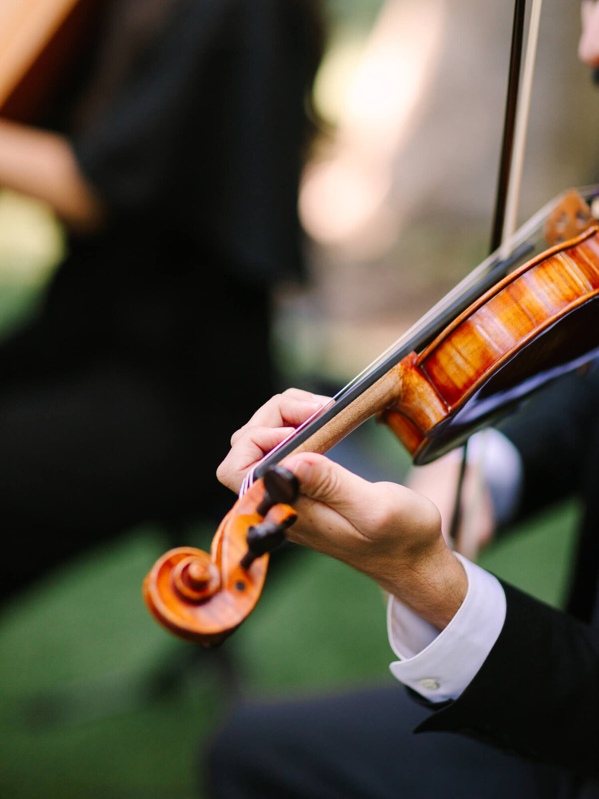 Close-up of a person in formal attire playing a violin outdoors, focusing on the right hand.