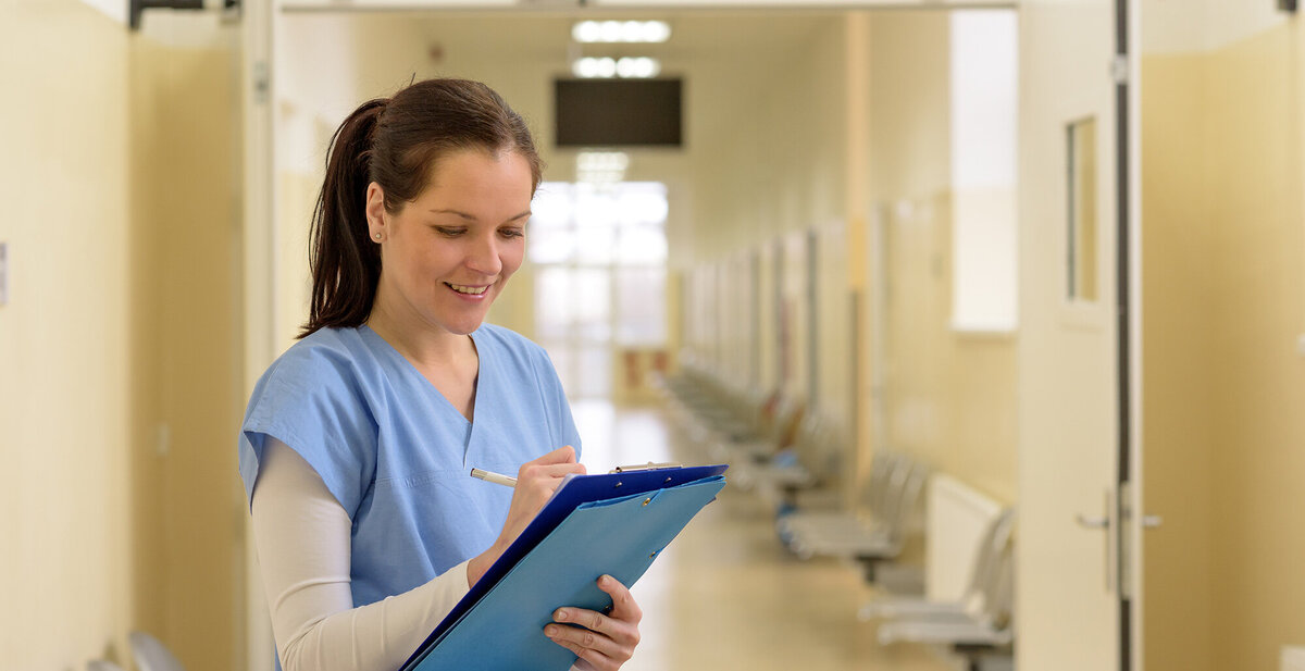 smiling-young-female-nurse-writing-in-patient-file-standing-alone-in-hospital-corridor