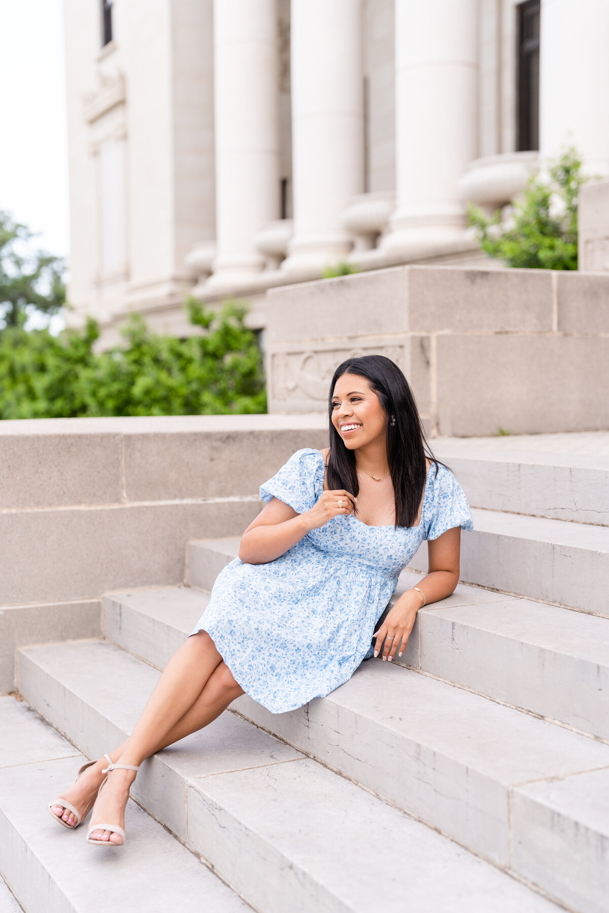 Texas A&M senior girl sitting and leaning back on stairs while touching hair and laughing away while wearing blue dress in front of the Administration Building