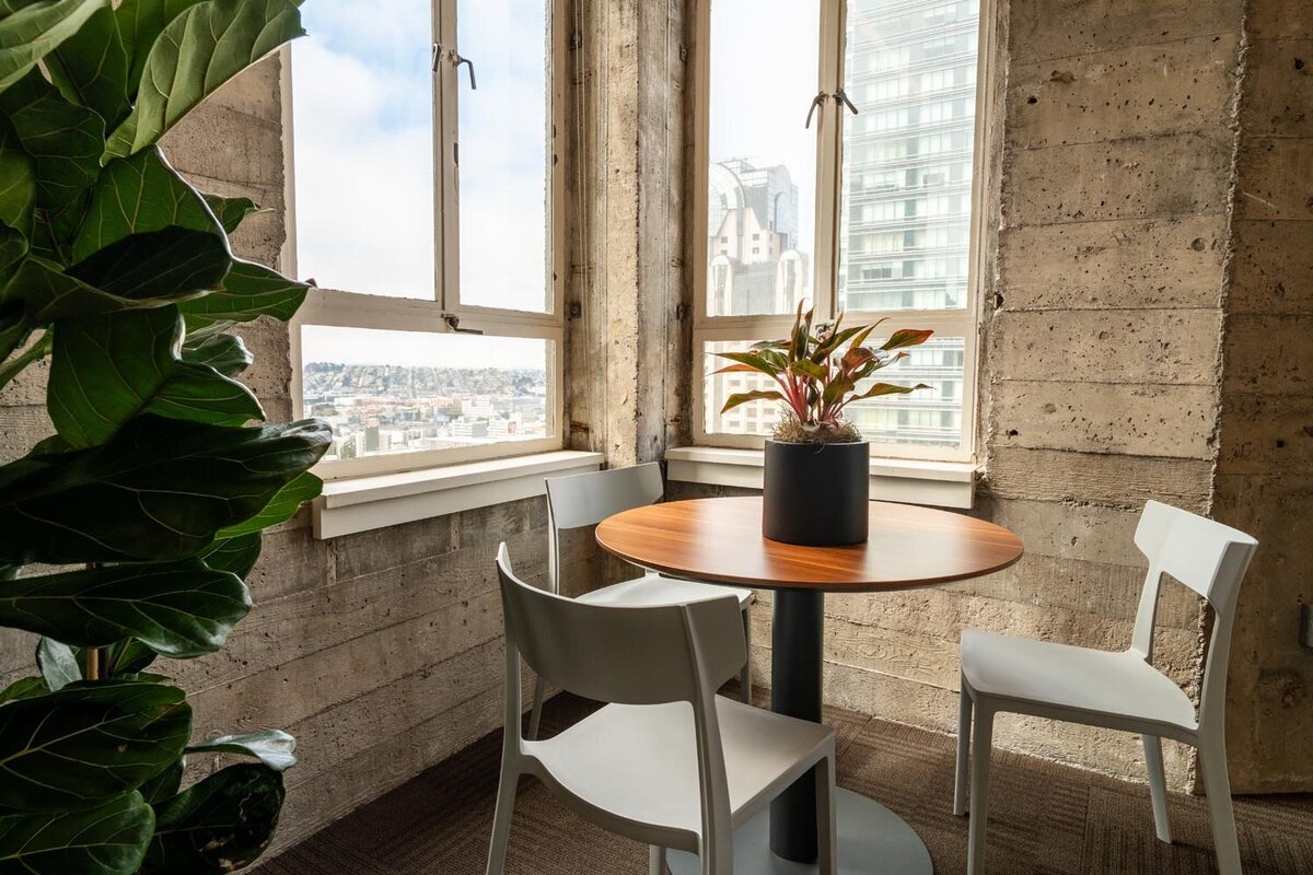 Small café-style seating area with a round wood table, light gray chairs, and a potted plant overlooking a panoramic city view.