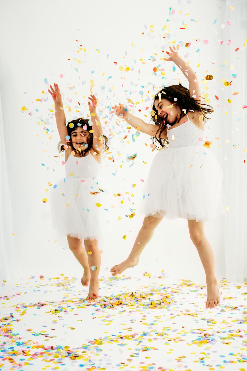 2 sisters jump as they throw confetti. they have white dresses and are on a white backdrop