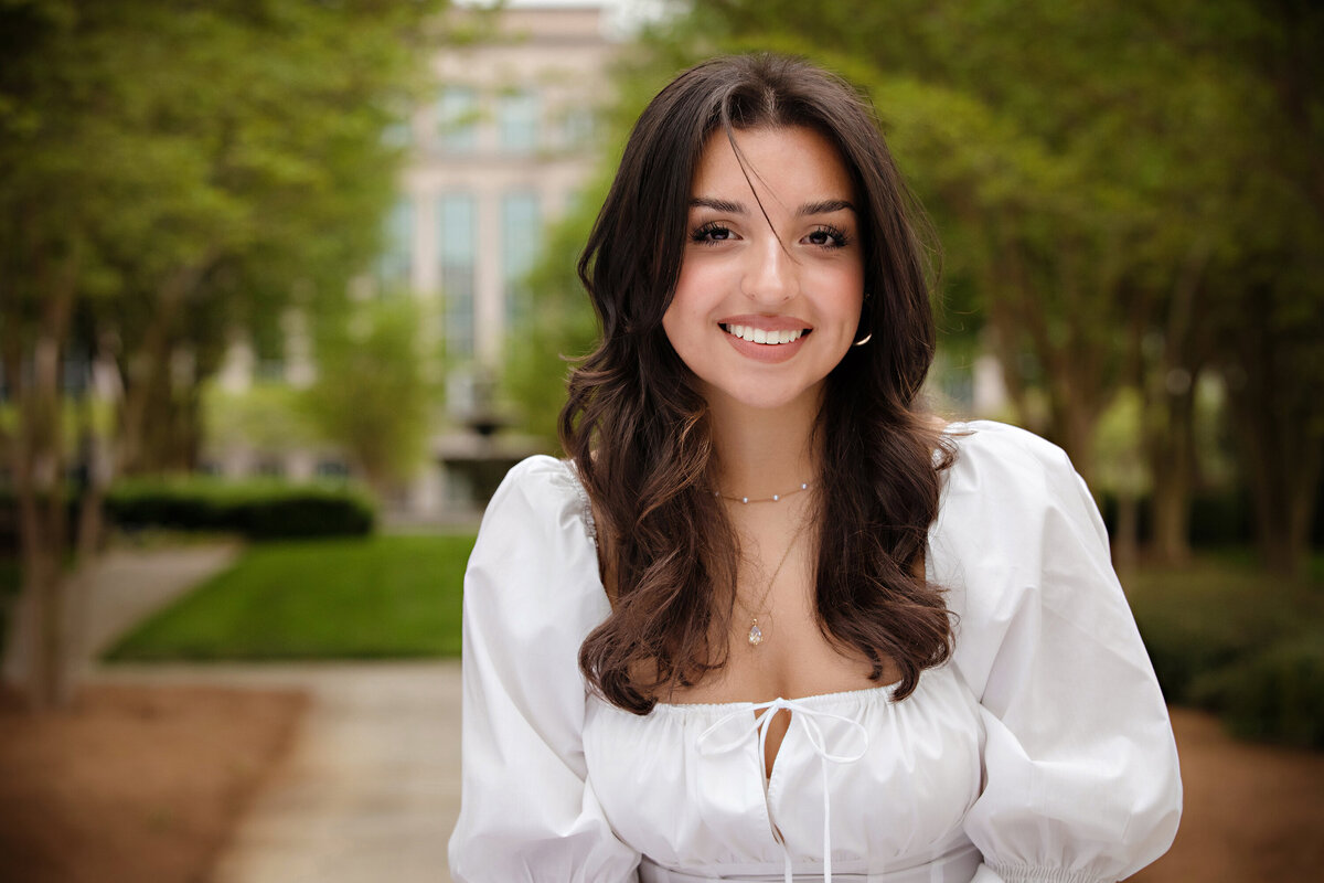Close-up senior portrait in white top with dark wavy hair, captured in a corporate park setting with fountain and courtyard backdrop.