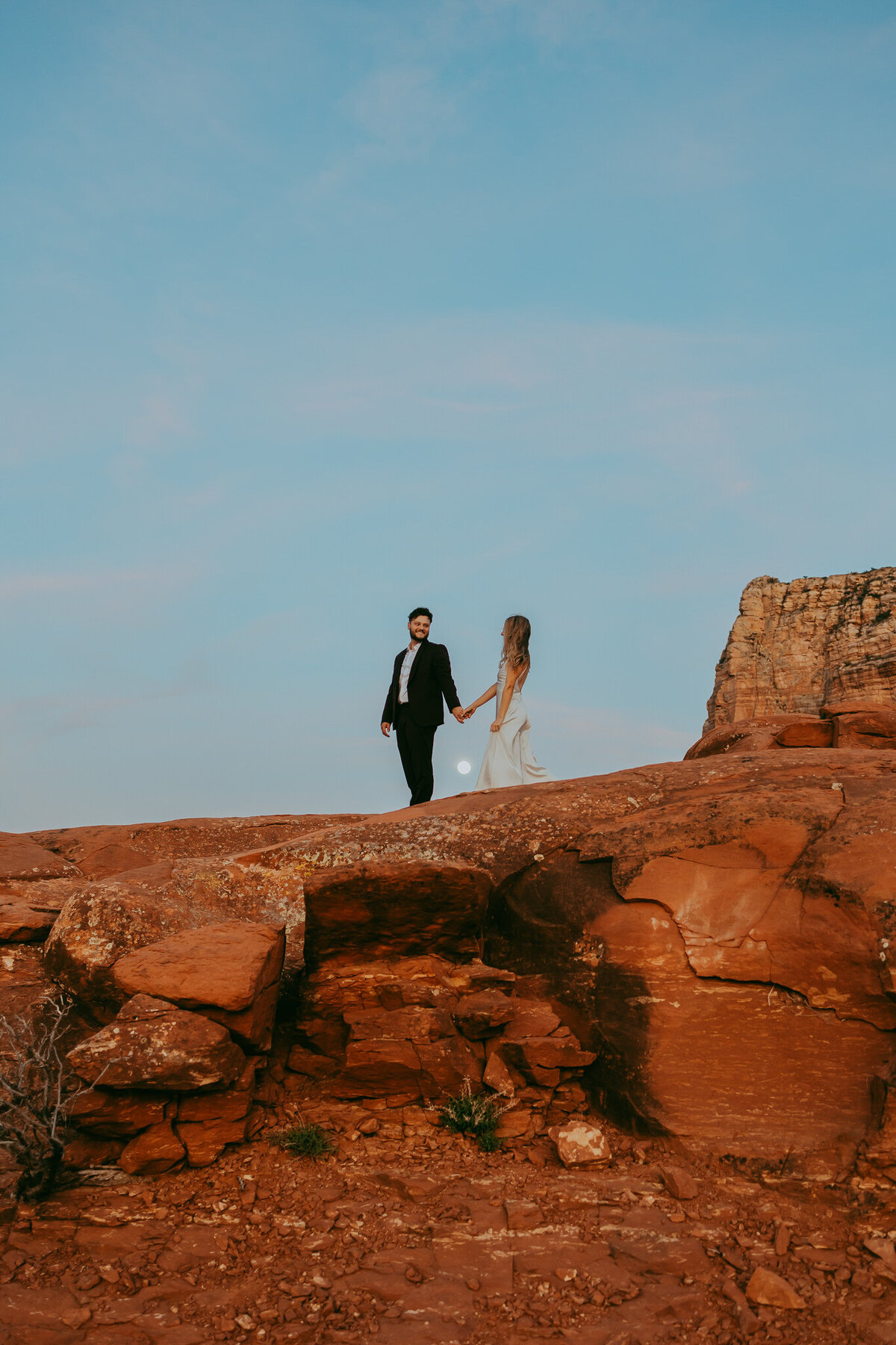 bride and groom hold hands on red rocks with full moon