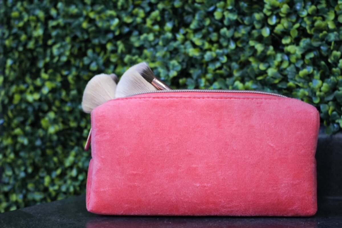 A pink makeup bag with makeup brushes in it, sitting in front of a grass wall