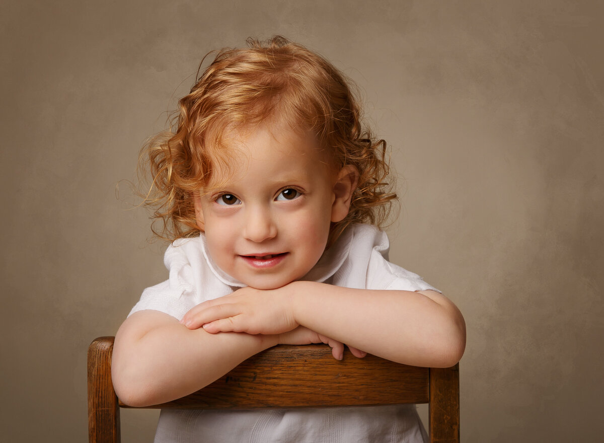 Little boy is sitting in a brooklyn ny photo studio for a family portraits. He is sitting on a backwards chair with his hands folded over the back and chin resting atop. He is smiling at the camera and has curly red hair. Captured by Chaya Bornstein, Brooklyn Family Photographer