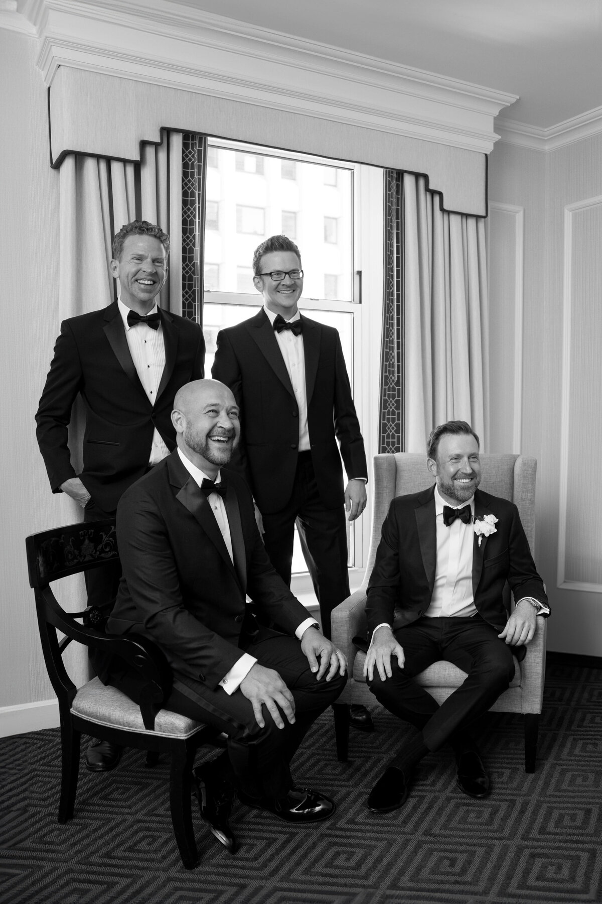 Four men in tuxedos are gathered in a room. Two are standing and two are seated, all smiling. The room has a window with a city view, curtains, and patterned carpet. It seems like a formal occasion or event.