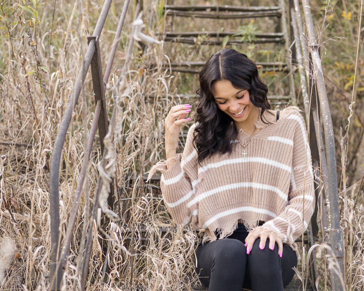 high school female senior, with long black hair, sitting on a set of stairs, looking down laughing, wearing black leggings, and a light tan and white stripped shirt
