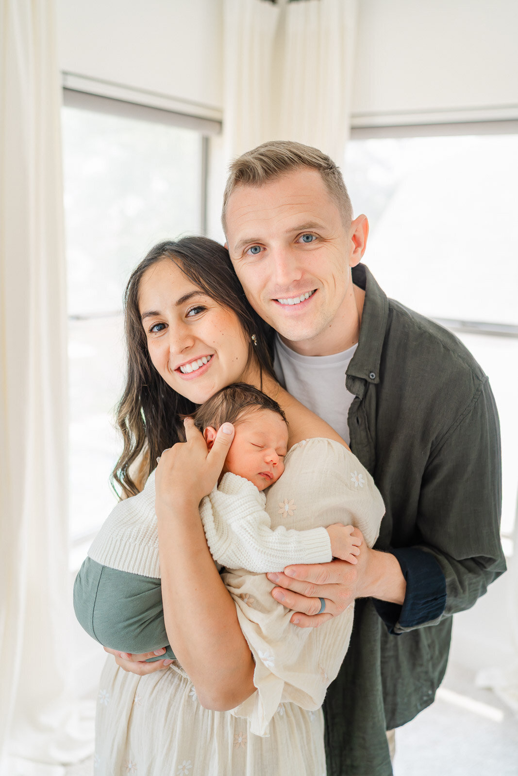 a mother and father stand in front of a window in their Round Rock home while holding their newborn.