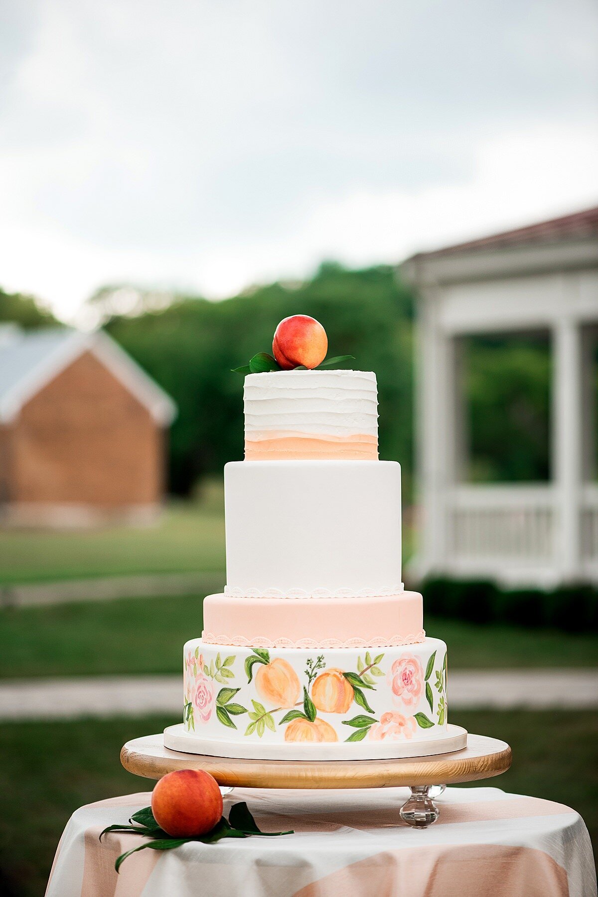 five tier hand painted water color peach themed wedding cake decorated with fresh peaches  and topped with a peach sitting on a table with a blush raw silk table cloth for a summer wedding at Ravenswood Mansion
