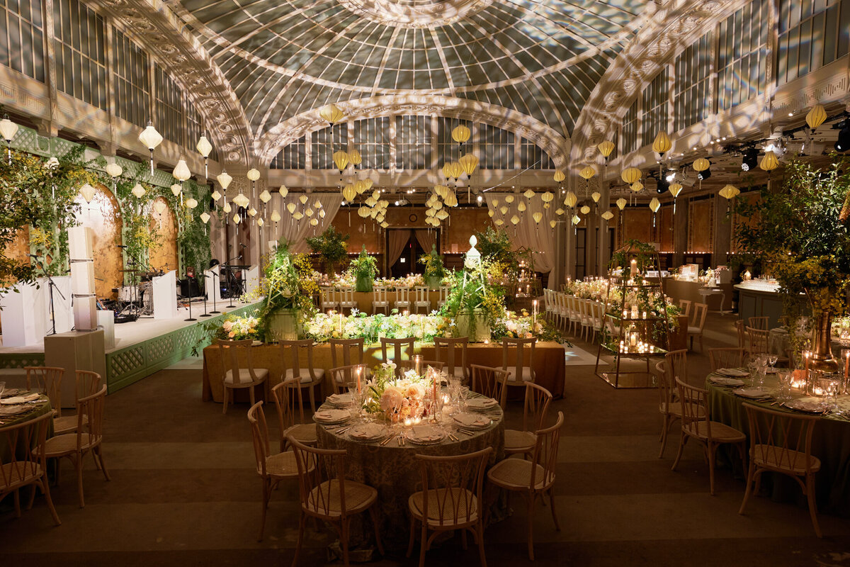 The photo was taken at the New York Public Library in New York City by the talented photographer Shawn Connell of Aaron Navak Films. It captures a grand indoor venue showcasing an elegant wedding reception setup. The round tables are beautifully adorned with floral centerpieces and candles, adding a touch of romance to the setting. The room boasts a high, glass-domed ceiling that enhances the space's grandeur. Additionally, string lights and hanging lanterns decorate the venue, casting a warm and enchanting ambiance over the whole scene.