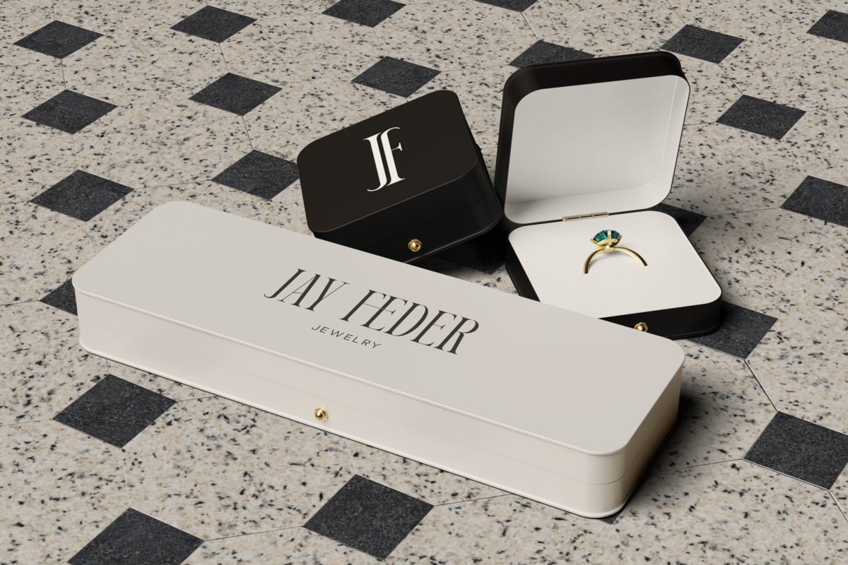 Jay Feder Jewelry packaging featuring a closed cream box, a black monogram box, and an open ring box displaying a gold ring with a green gemstone on a tiled floor.