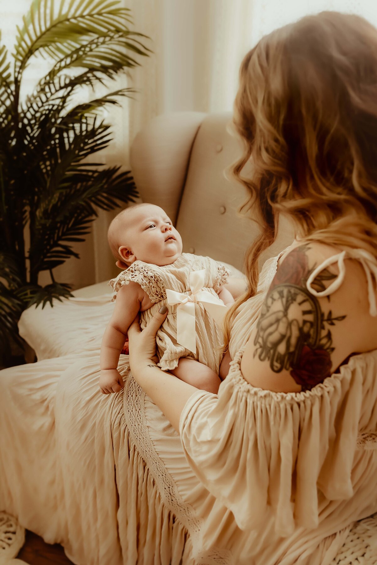 A woman holding a baby dressed in white, sitting in a softly-lit room with a plant in the background. The woman has long wavy hair and a tattoo on her right arm.