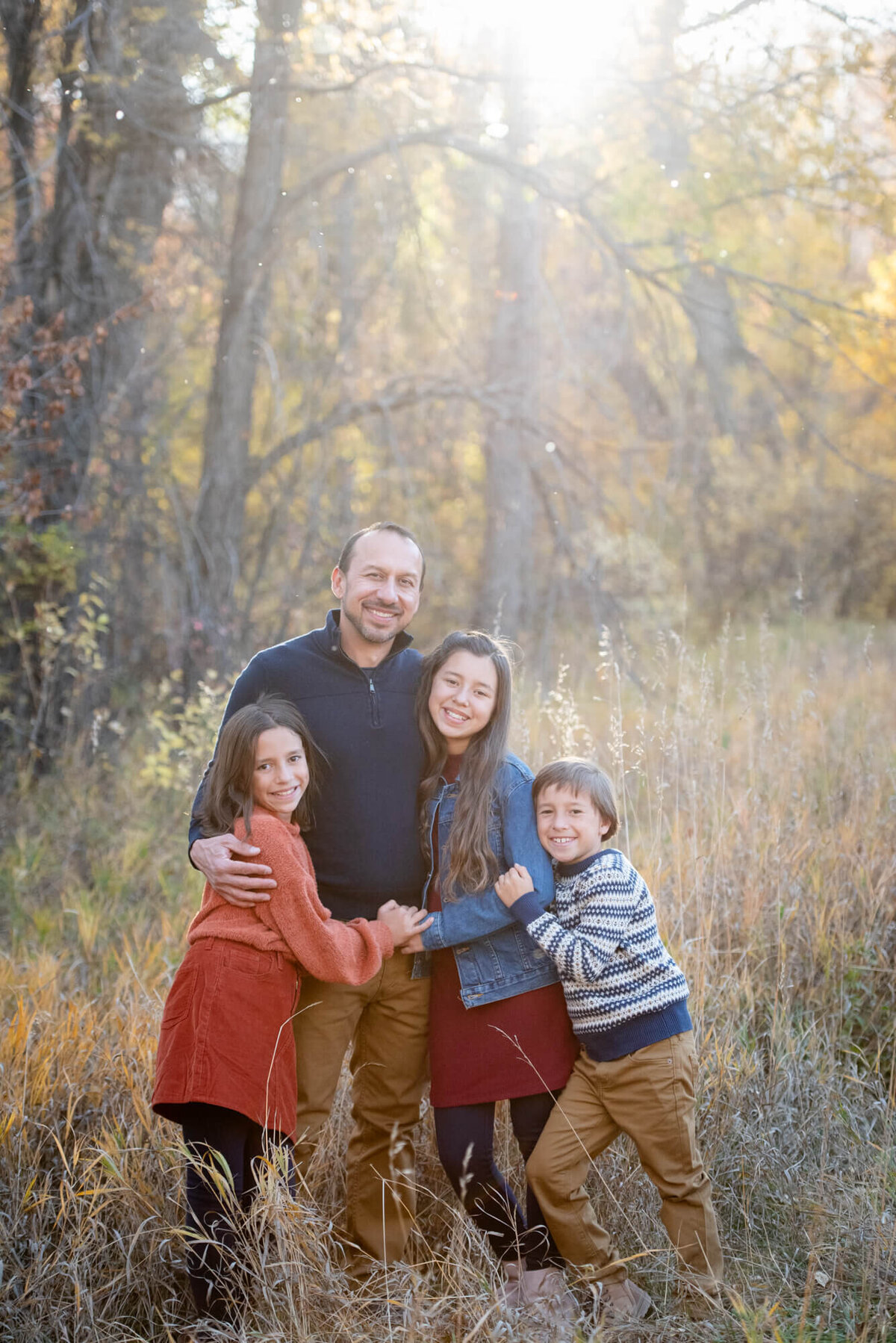 A father in a blue sweater  is hugged y his three young children in a forest at sunset