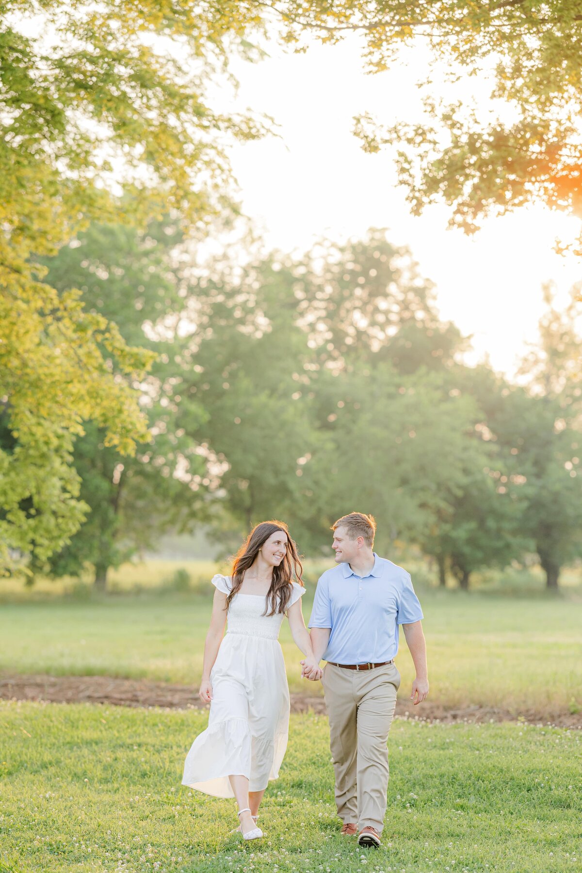 Newly engaged couple walks in a field at sunset holding hands and smiling at each other