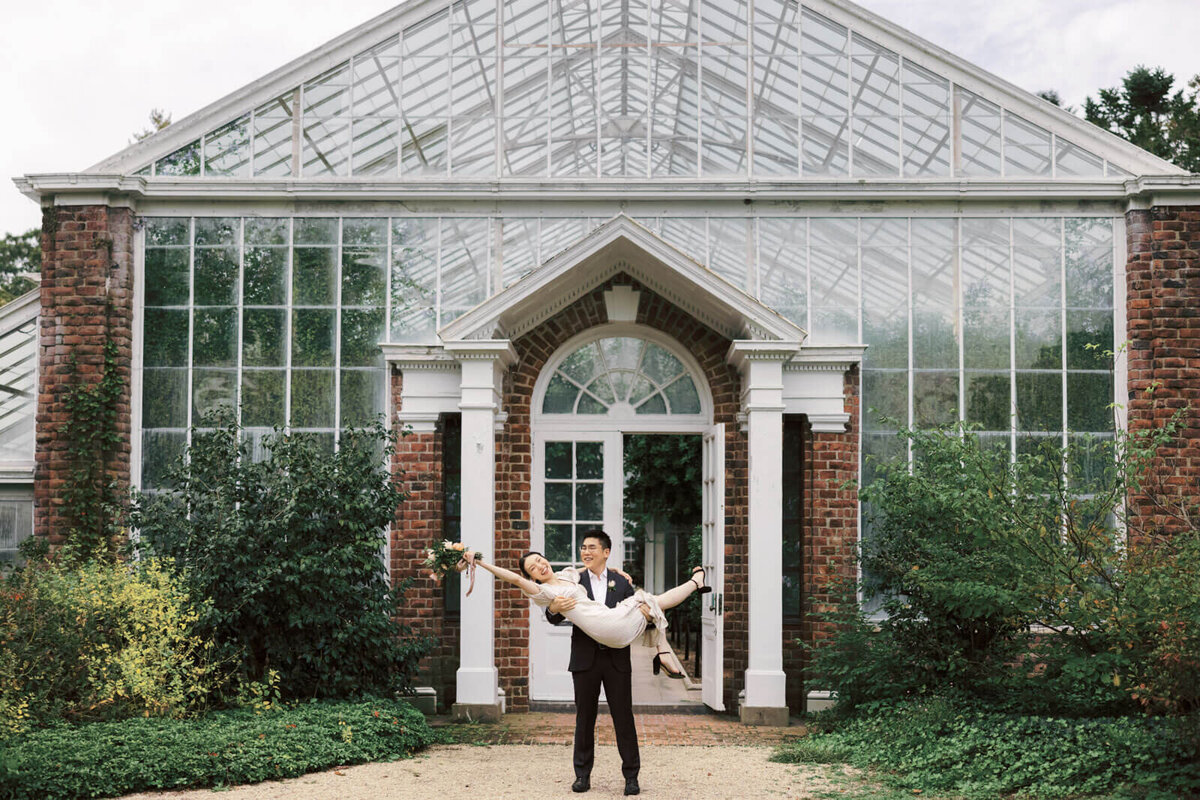 The engaged man is carrying his fiance in front of the greenhouse at Coe Hall at Planting Fields Arboretum, NY. Image by Jenny Fu Studio