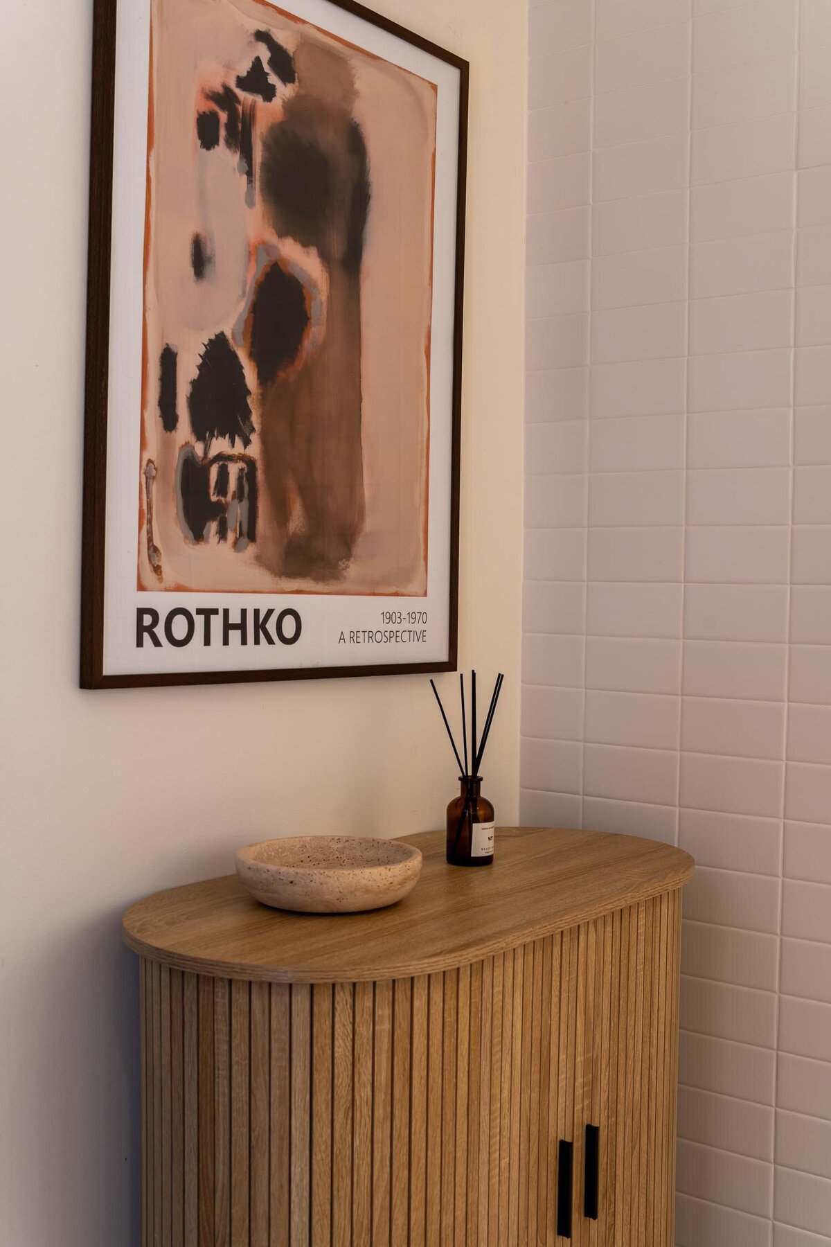 Styled console featuring a framed Rothko print, ribbed wood cabinet, ceramic bowl, and diffuser bottle against a tiled wall.