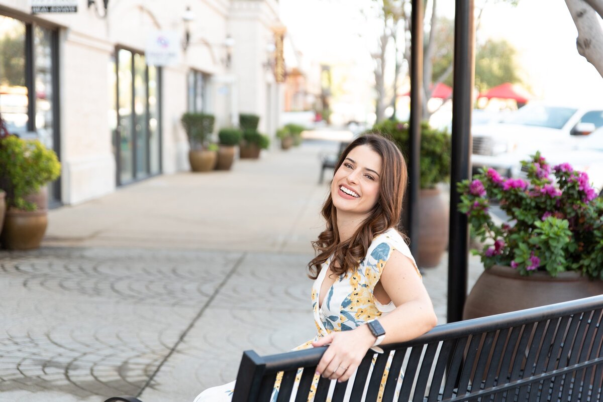 Woman smiling sitting on a black bench along an outdoor storefront walkway.