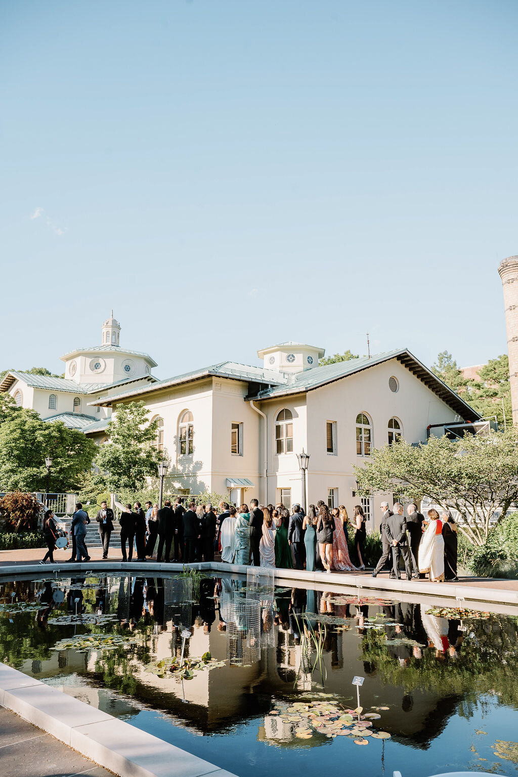 Rhode Island Wedding Photographers | A large group of people in formal attire gather outside a grand building with a lush garden and reflection pool, under a clear blue sky.