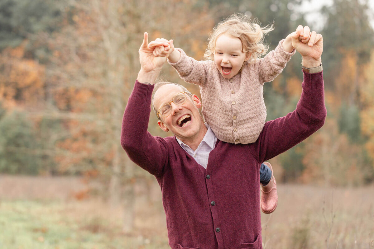 Dad in burgundy sweater holding his daughter up over his shoulder and flying her around like an airplane. They both have huge smiles on their faces. There is a tree line behind them in beautiful fall hues.