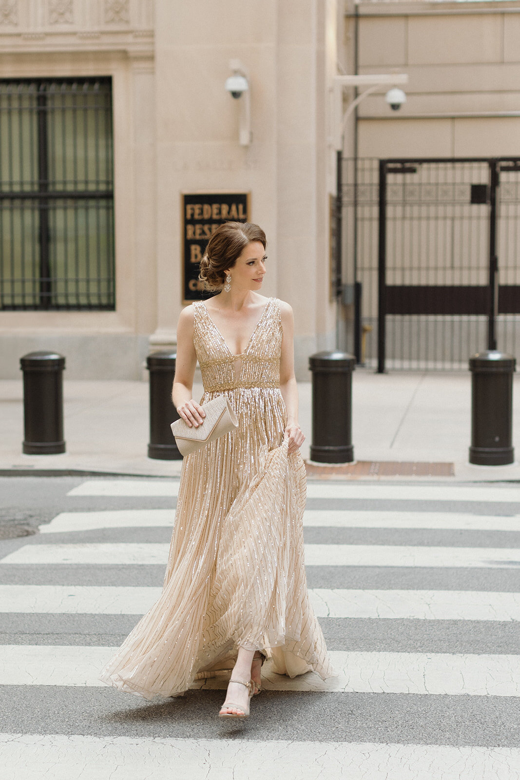 woman walking in crosswalk in gold sparkly gown and holding a clutch. She is holding up her gown and looking off to the side.