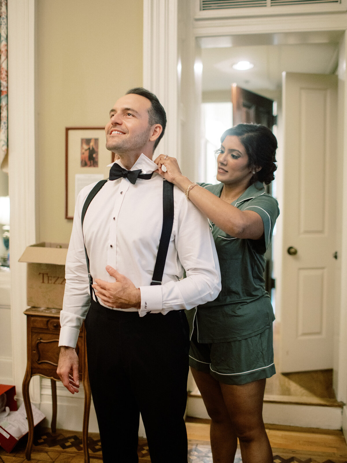A joyful groom with a professionally styled bowtie, showcasing a special moment as the bride helps him prepare, highlighting love and partnership on their wedding day.