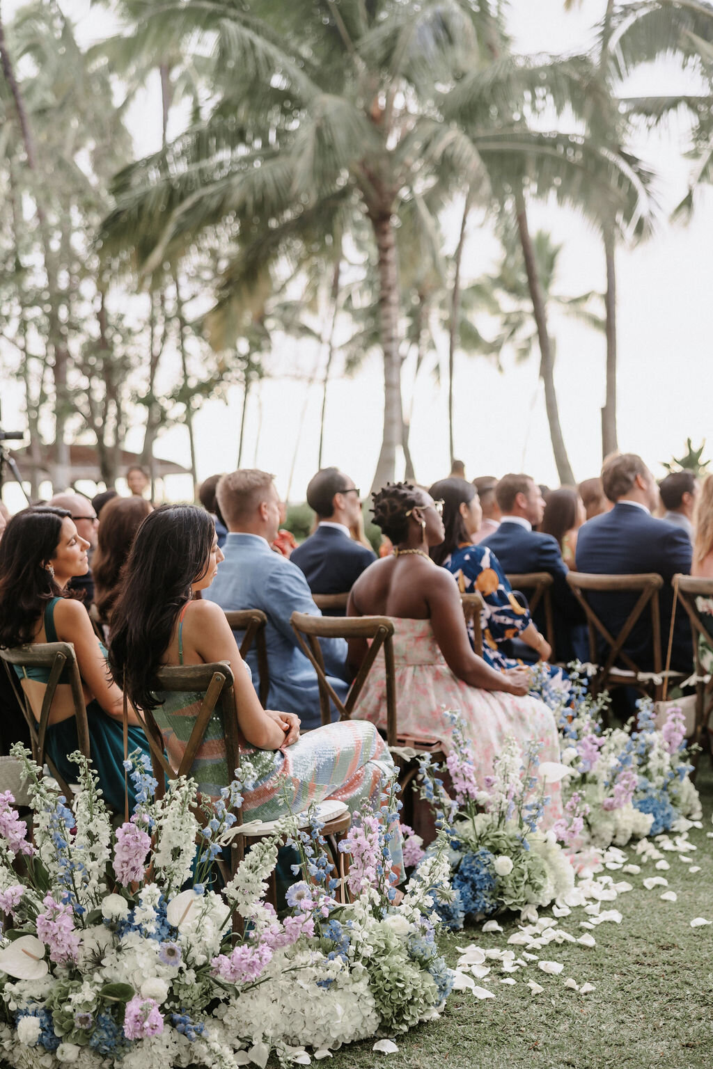 wedding guests seated in multicolor light spring colors with aisle florals that are purple, white and blue and palm trees in the distance at lanikuhonua