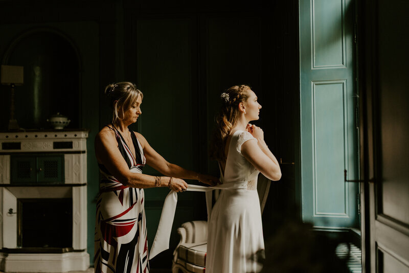 Maman ajustant la robe de mariée de sa fille face à une fenêtre lors d'une séance photo mariage en Vendée.