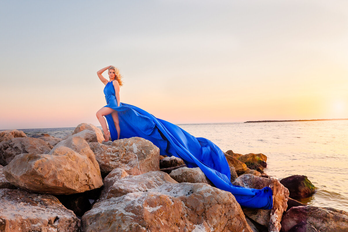 Girl in a blue flying dress posing barefoot on beach rocks at sunset, with the dress trailing dramatically behind her by the ocean.