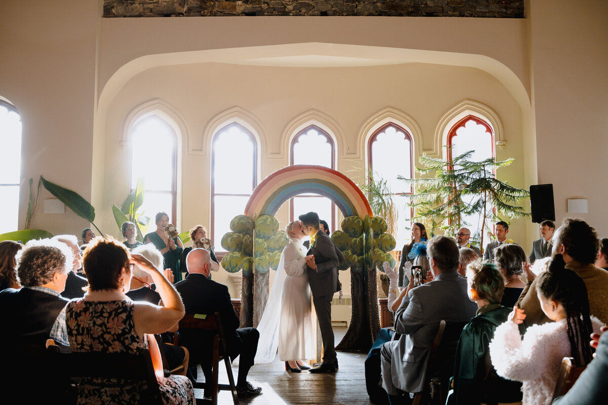 A couple during their first kiss at their wedding while all of their guests cheer.