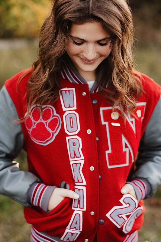 senior girl wearing red letterman jacket in golden field at sunset