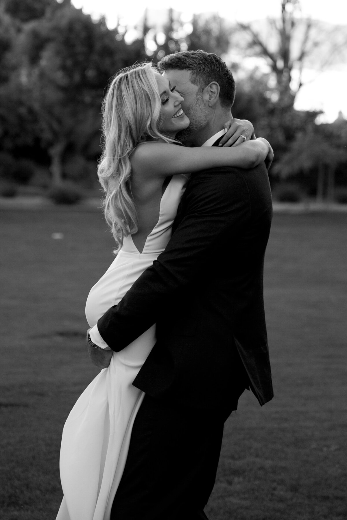 Groom picking up a bride under the butt and lifting her up. Bride has a soft smile on her face as her groom kisses her neck.