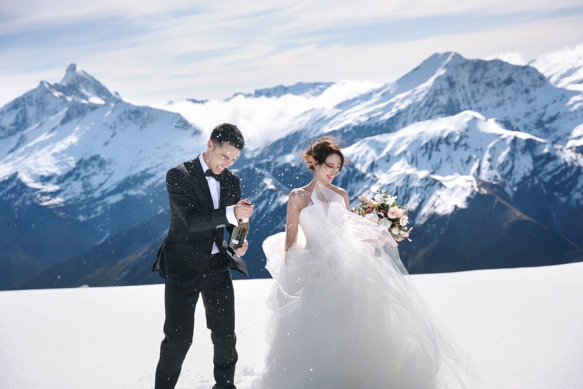 A happy couple popping champagne on a helicopter photoshoot in the snowy mountains.