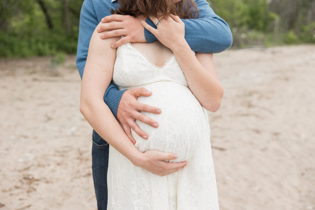 Expectant couple holding each other on a beach and looking out over the lake