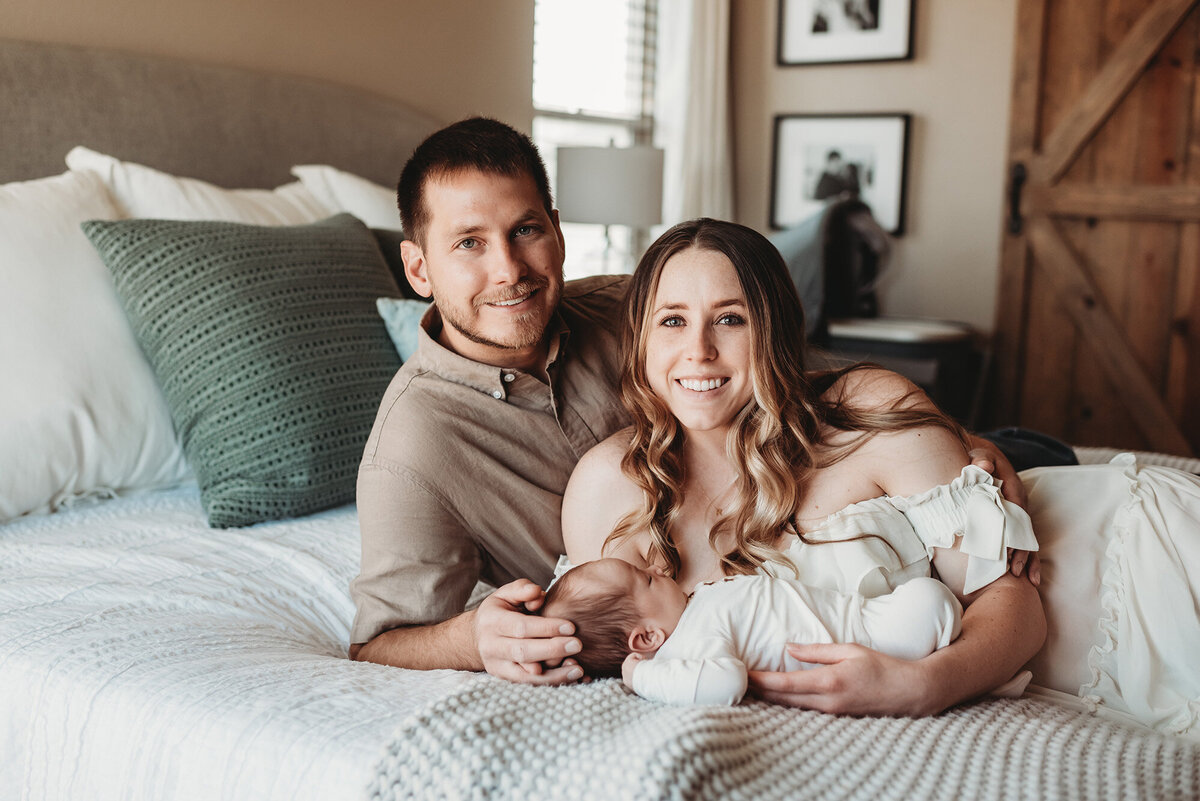 parents laying on bed holding newborn baby while they look at Denver newborn photographer