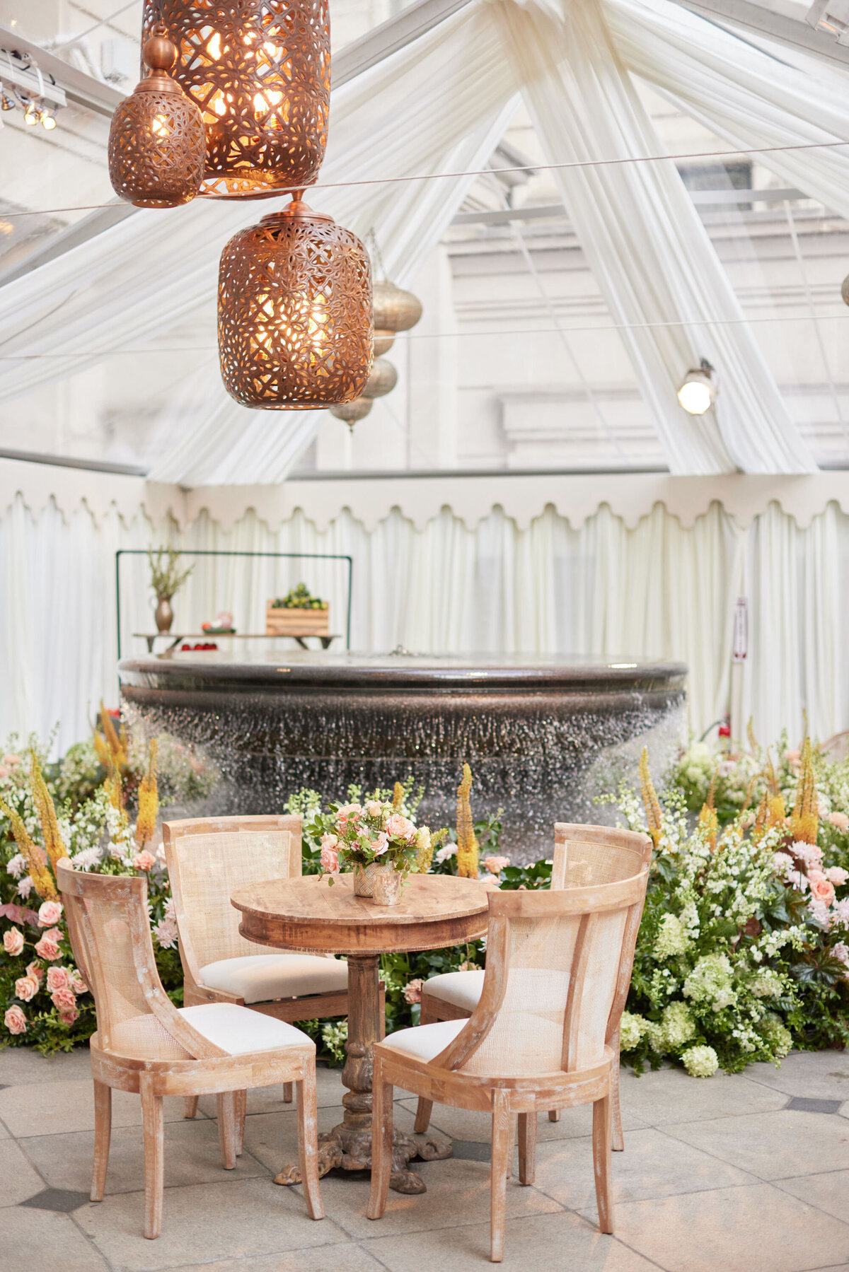 The photo was taken at The Willard Intercontinental in Washington, D.C., a top wedding venue and hotel. The videographers from Clayton Films captured the cozy outdoor dining setup beautifully. The scene includes a round wooden table, four beige chairs, and charming decor with flowers and greenery. Intricately designed wicker lanterns hanging above the table add a soft and inviting glow to the space, making it truly enchanting.