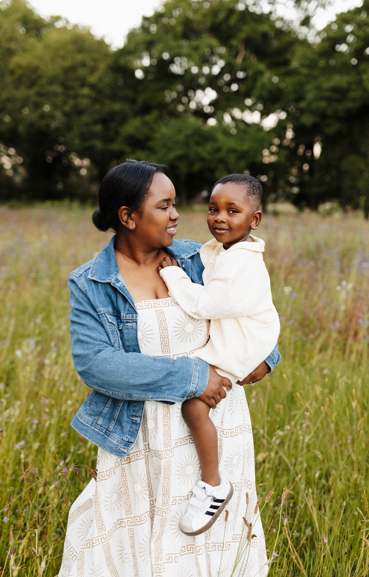 A mother wearing a denim jacked holds her son in a field of grass at sunset during a family photoshoot in canterbury