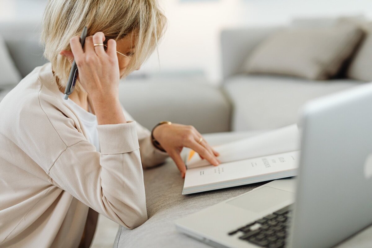 Femme blonde, gilet beige, travaillant sur son cahier posé sur un canpé, ordinateur au premier plan.