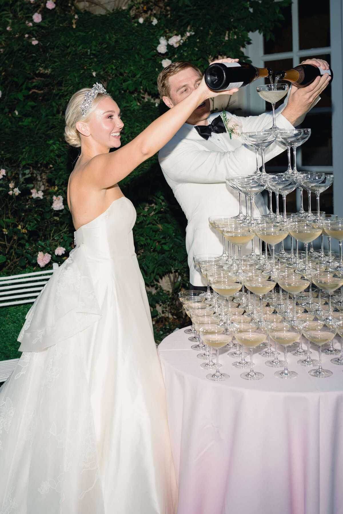 bride and groom pour champagne into their champagne tower which is set outside in the gardens of euridge manor on pink linen for their romantic wedding