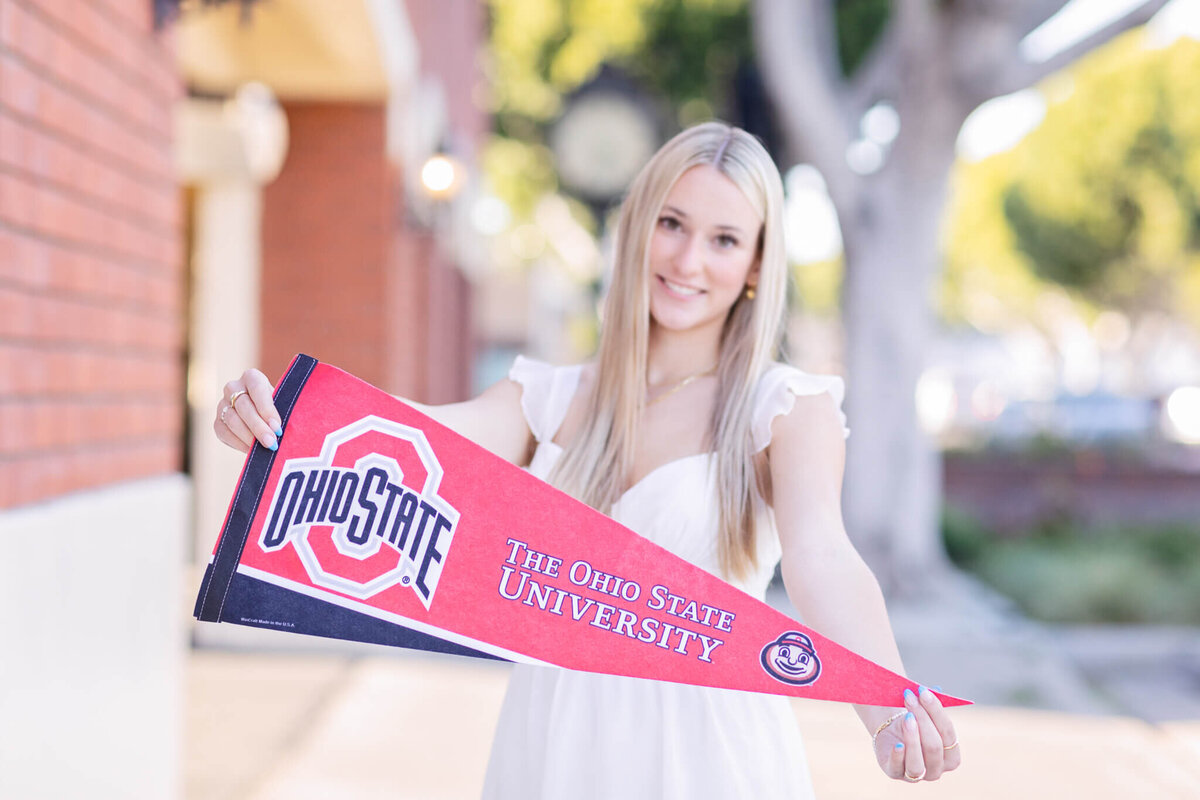 A woman in a white dress holds a red pennant that reads The Ohio State University with the university logo. She stands outdoors with blurred trees and a brick building in the background.