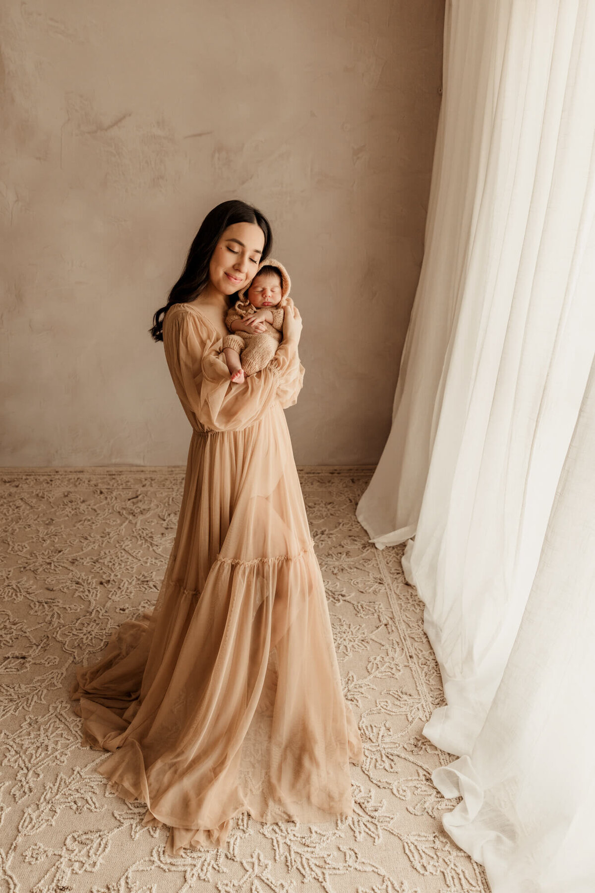 Mother holds her son next to her cheek for a newborn photo session in Oklahoma City while wearing a tan sheer dress.