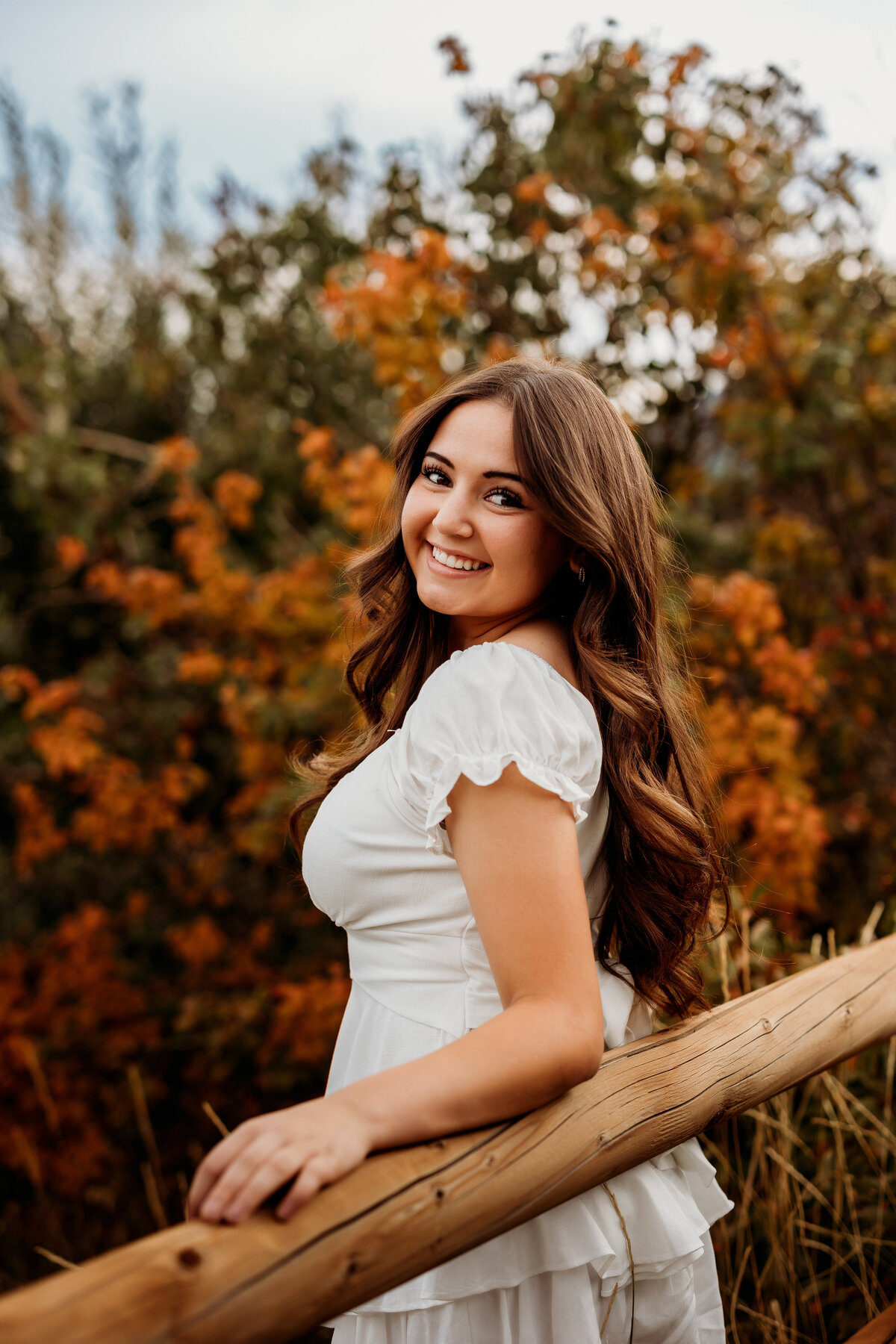 Girl poses with her back against a fence and fall trees behind her while wearing a white dress for her high school senior photos