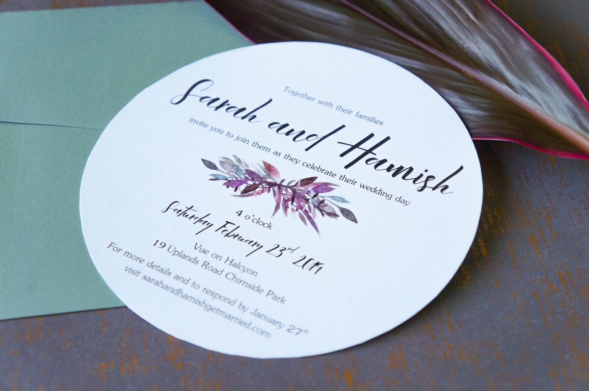 Round wedding invitation with purple foliage and a fun font, sitting on a sage green envelope
