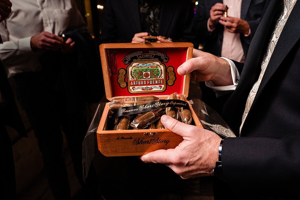 A groom wearing a black tuxedo and white shirt passes out Cuban cigars in a small wooden cigar box with red lining to other wedding guests.