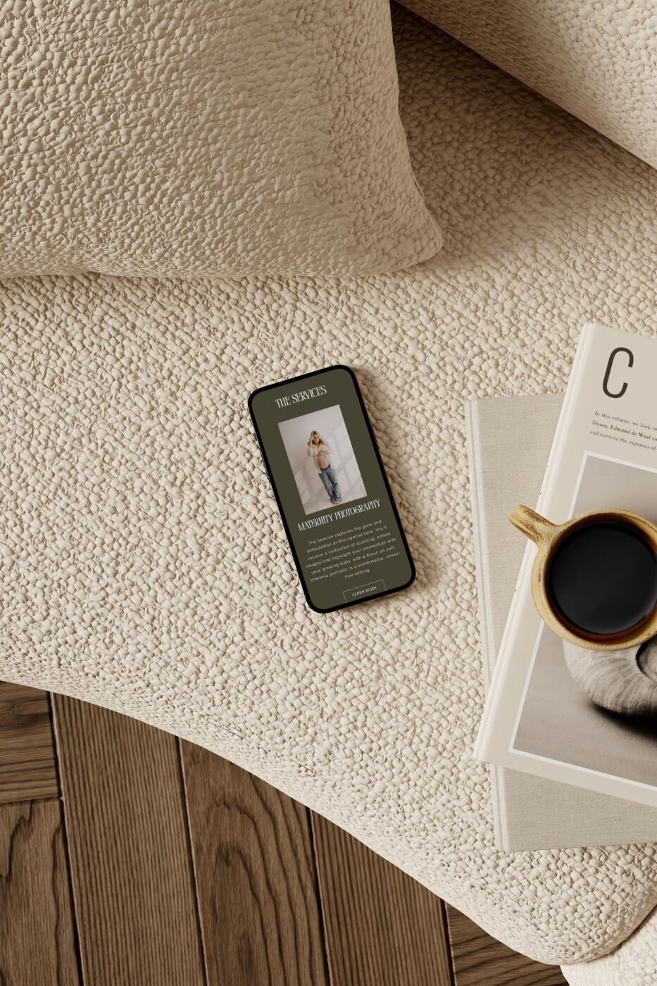 Smartphone resting on a textured beige sofa beside a magazine and cup of black coffee, displaying a maternity photography services page with a pregnant woman posing in casual jeans and a cream top.