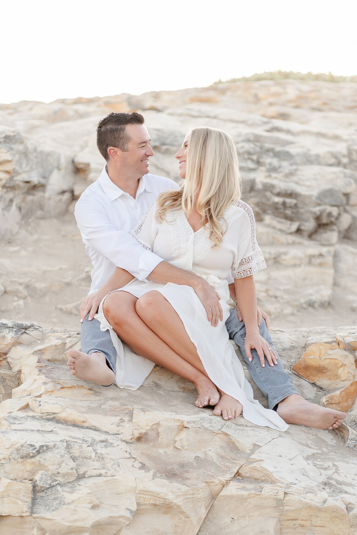 husband and wife wearing white and sitting together on a tock at the beach