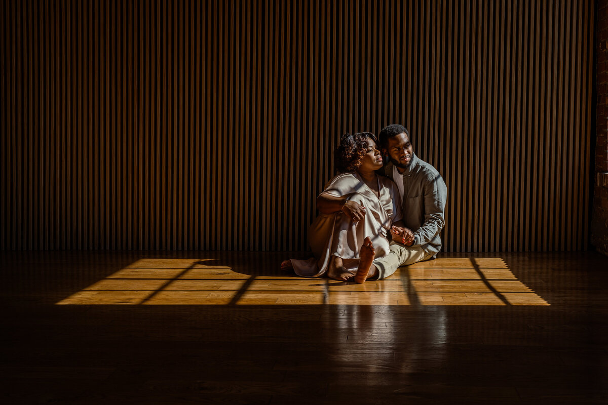 A sophisticated portrait of parents sitting together on a sunlit wooden floor, surrounded by warm, natural light and dramatic shadows during a family session in NYC. The minimalistic and intimate composition embodies the depth of connection and legacy.