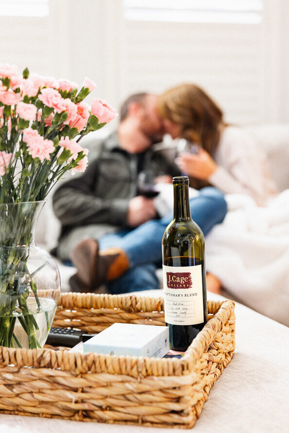 Wine bottle in a basket on the table with couple kissing in the background.