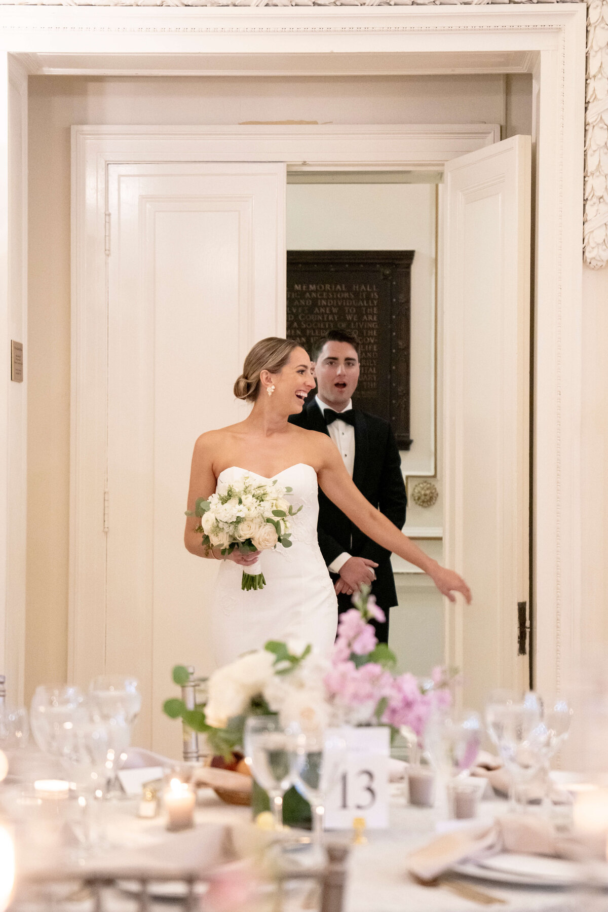 A bride in a strapless white gown holds a bouquet as she enters a room, followed by a groom in a tuxedo. The table in the foreground is set for a wedding reception with floral arrangements and candles.