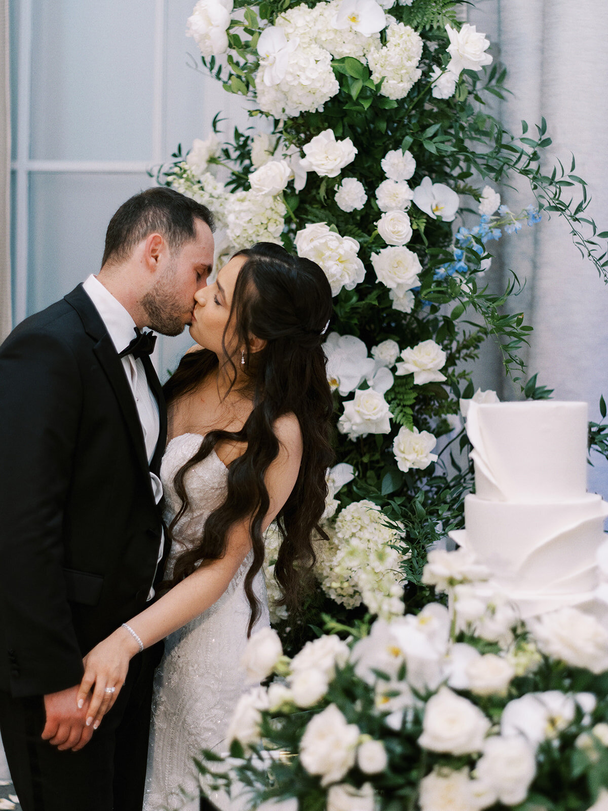 A couple in wedding attire shares a kiss next to a white, tiered wedding cake and lush floral arrangements featuring white flowers and greenery at their classic Calgary wedding.